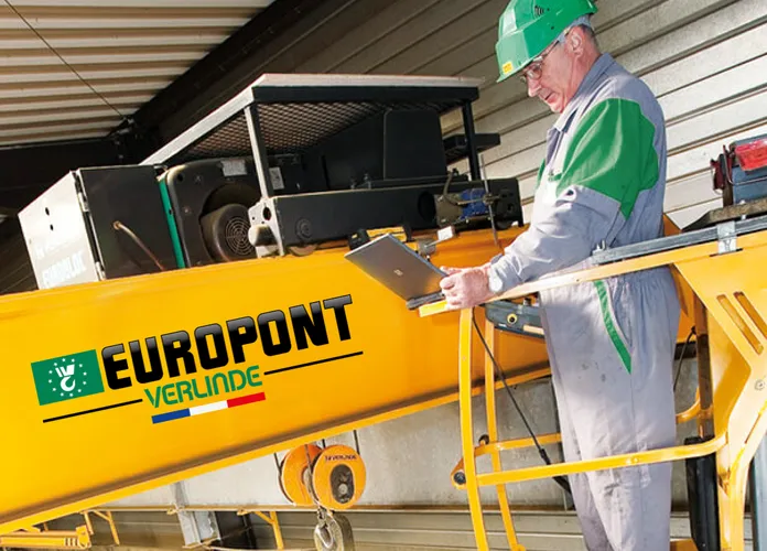 Technician in safety gear works on a yellow Europont Verlinde crane in a warehouse, checking controls with a laptop.