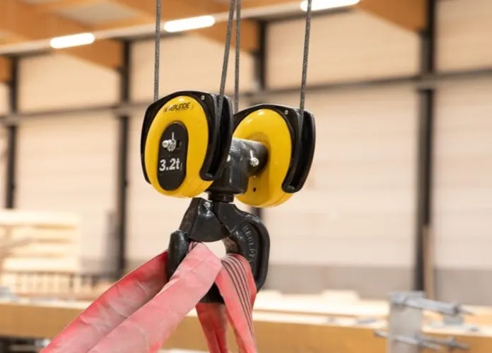 A yellow crane hook lifting a wooden beam with red straps in an industrial setting. The hook is marked with a 3.2-ton capacity.